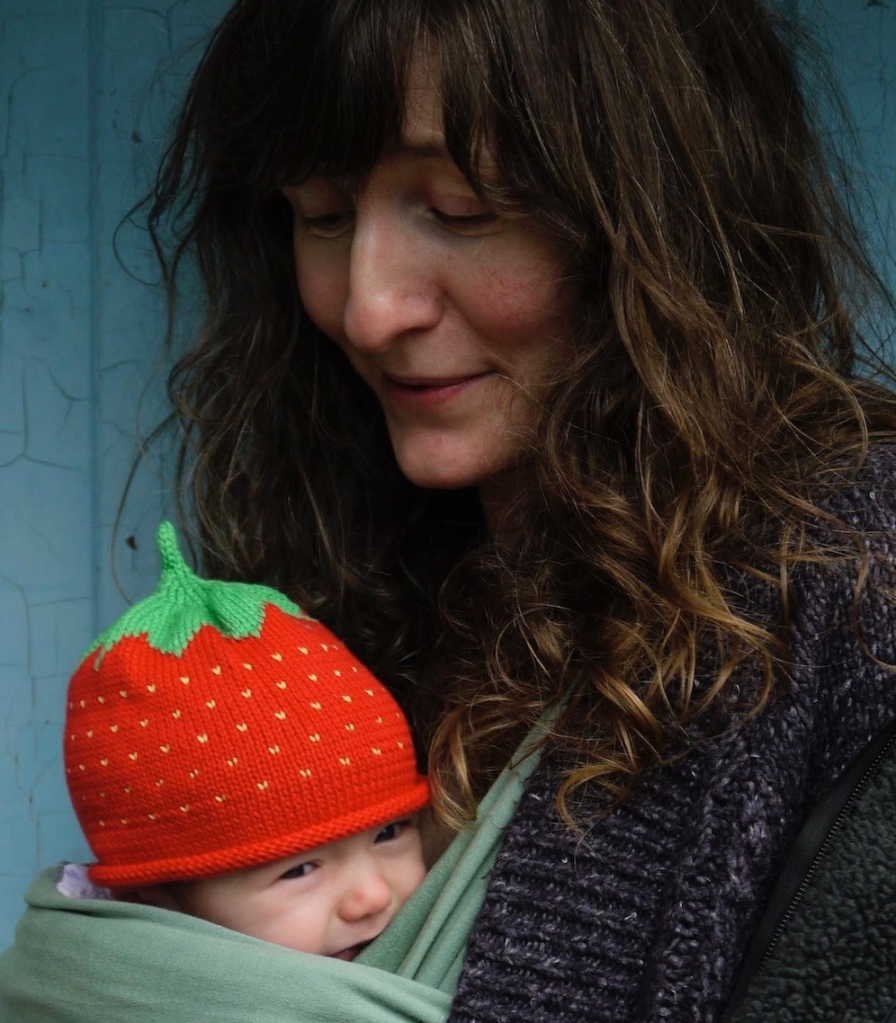 Ali with long flowing brown hair and her baby in a sling with a strawberry hat