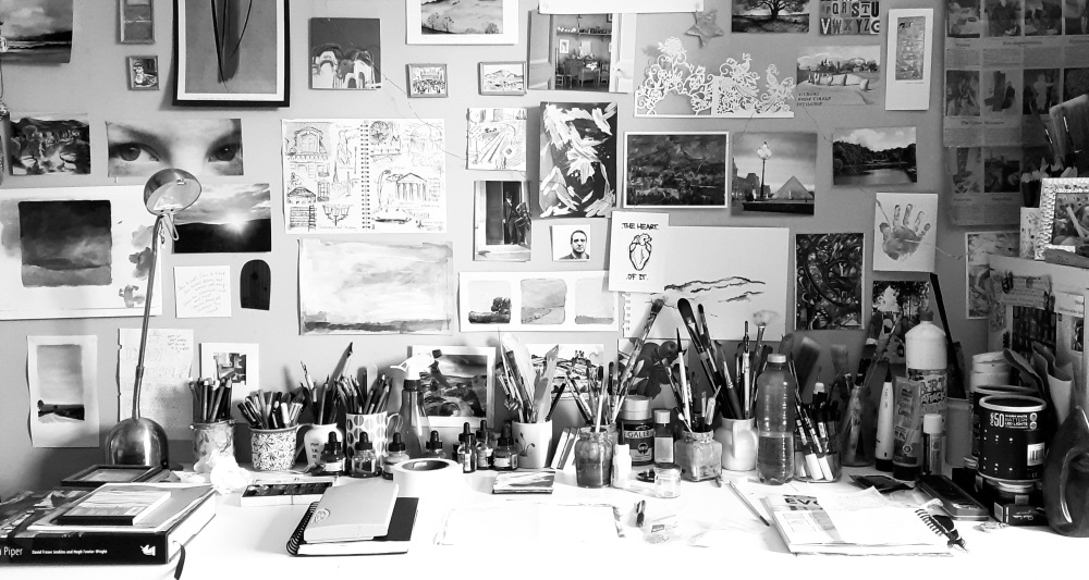 A black and white photo of an artist's desk with several paintings and paint brushes