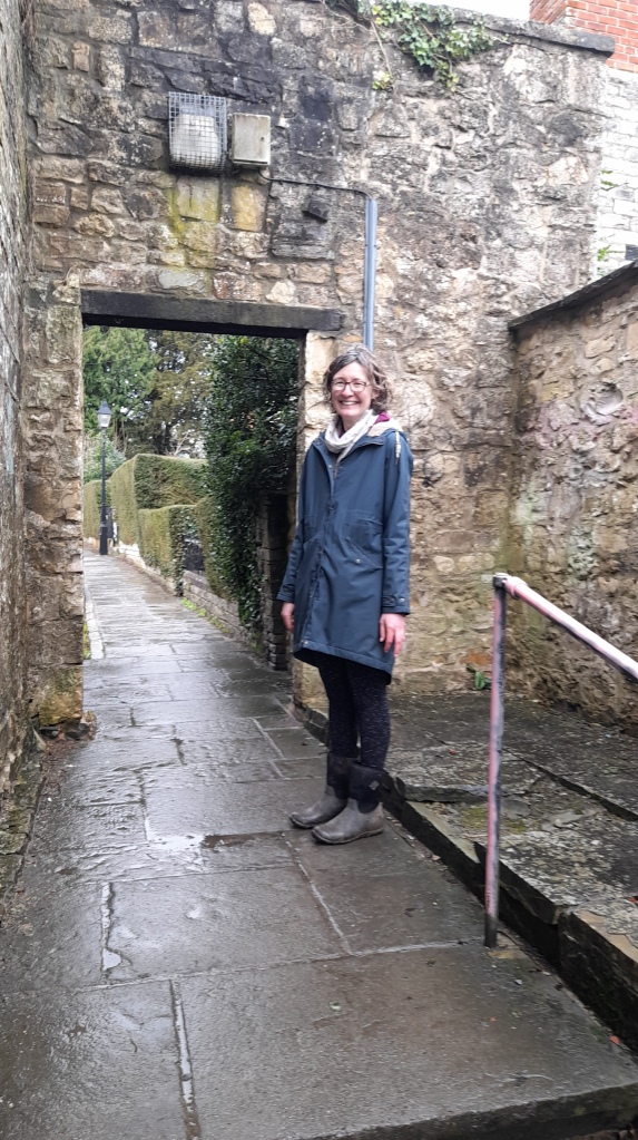 Ali stands by a stone doorway she is wearing a dark blue coat, slim with dark hair