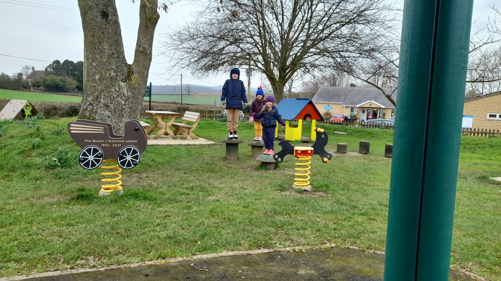 A playpark with three kids standing on tree stumps looking rather like meerkats amongst the colourful play equipment and daffodils