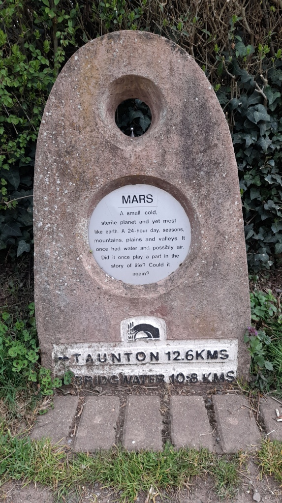 A marker stone with a small metal planet. It reads: Mars. A small, cold sterile planet and yet most like earth. It has 24 hour days, seasons, plains and valleys. It one had water and possibly air. Did it once play a part in the story of life? Could it again? Taunton 12.6 kms Bridgwater 10.8 kms.