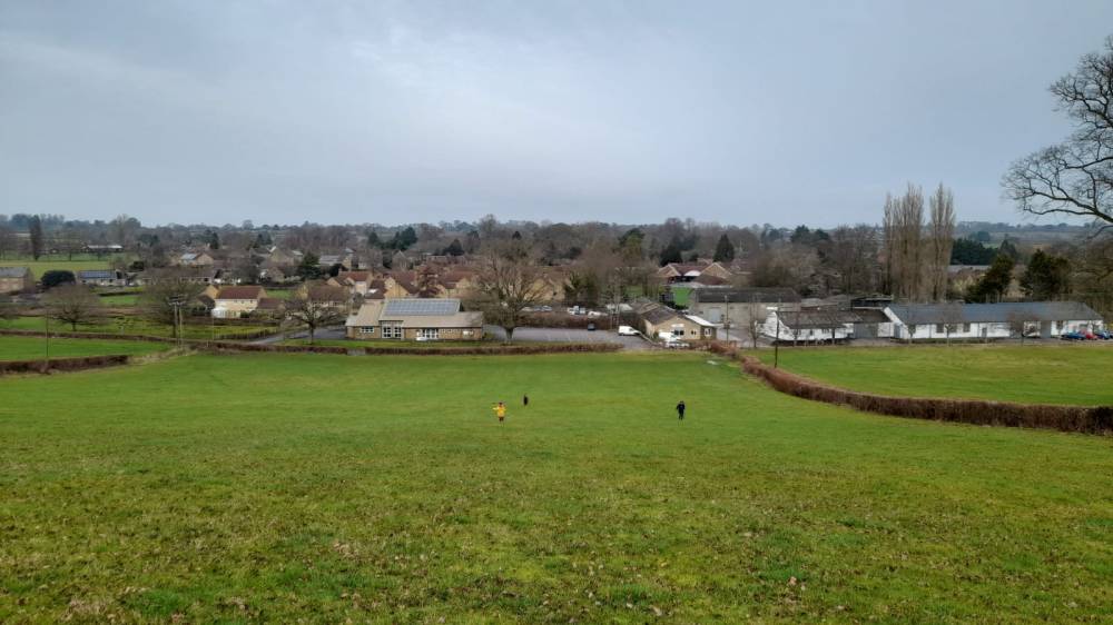Running down that hill! 3 tiny figures running down a field of green with village hall and buildings the destination