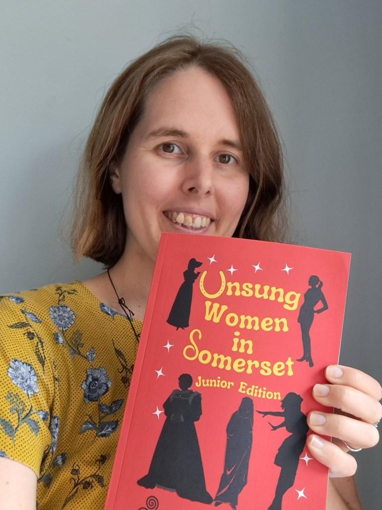 Helen is holding her book Unsung women in Somerset. Helen has hazel eyes and short mid brown hair, is smiling and wearing a mustard coloured floral top.